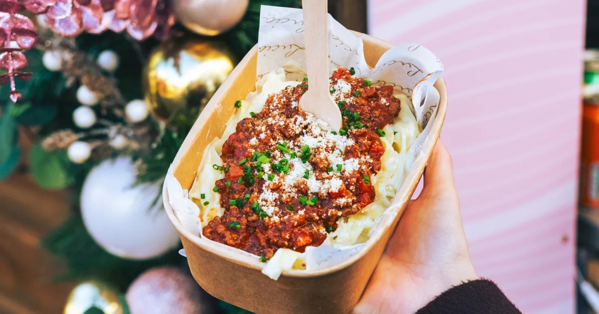 Person holding a cardboard container of loaded fries topped with red sauce, crumbled white cheese, and green herbs, with festive Christmas decorations blurred in the background