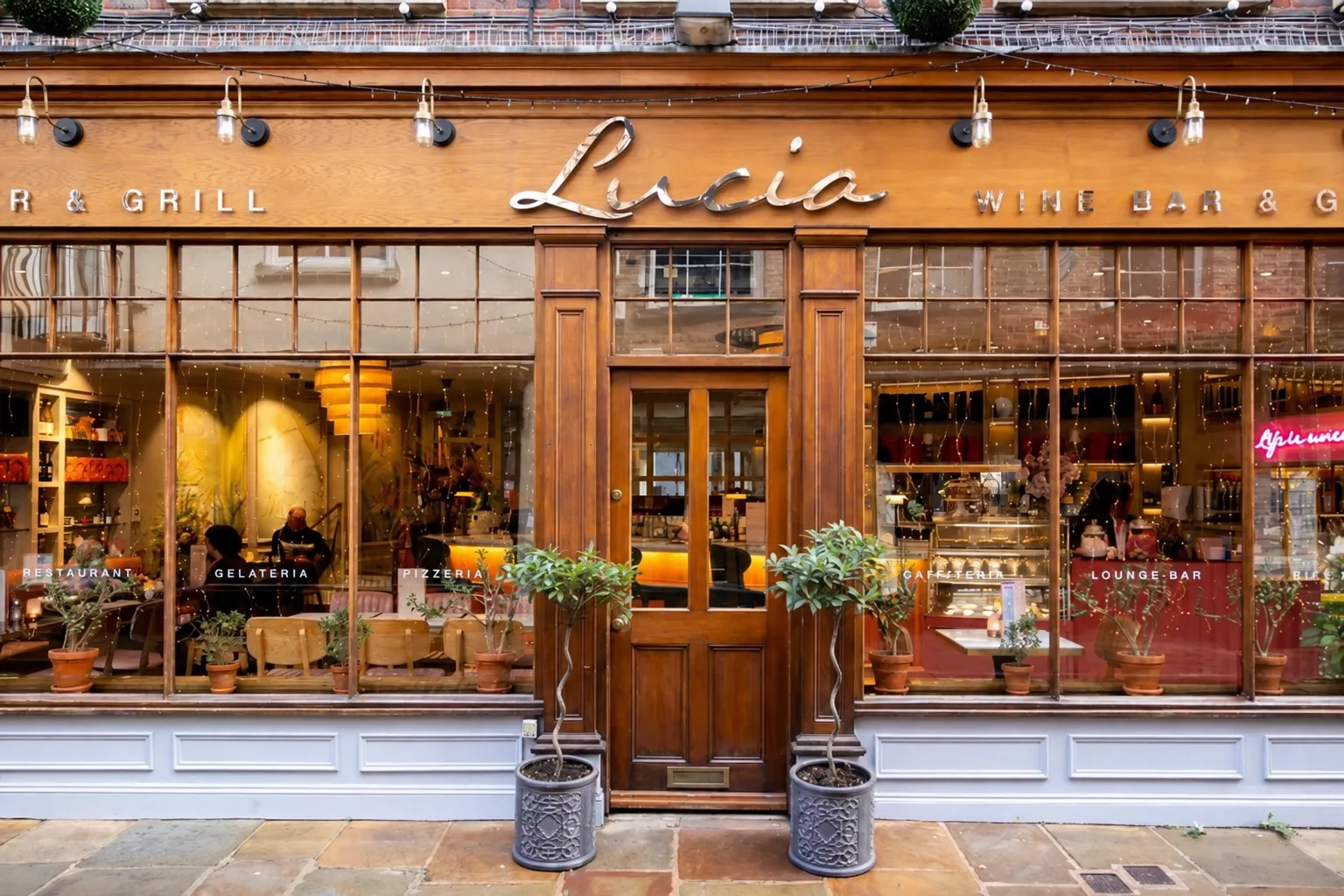 Storefront of Lucia restaurant with wooden facade, large windows displaying interior seating areas, silver signage, potted plants flanking the entrance, and pendant lights strung across the upper section of the building.