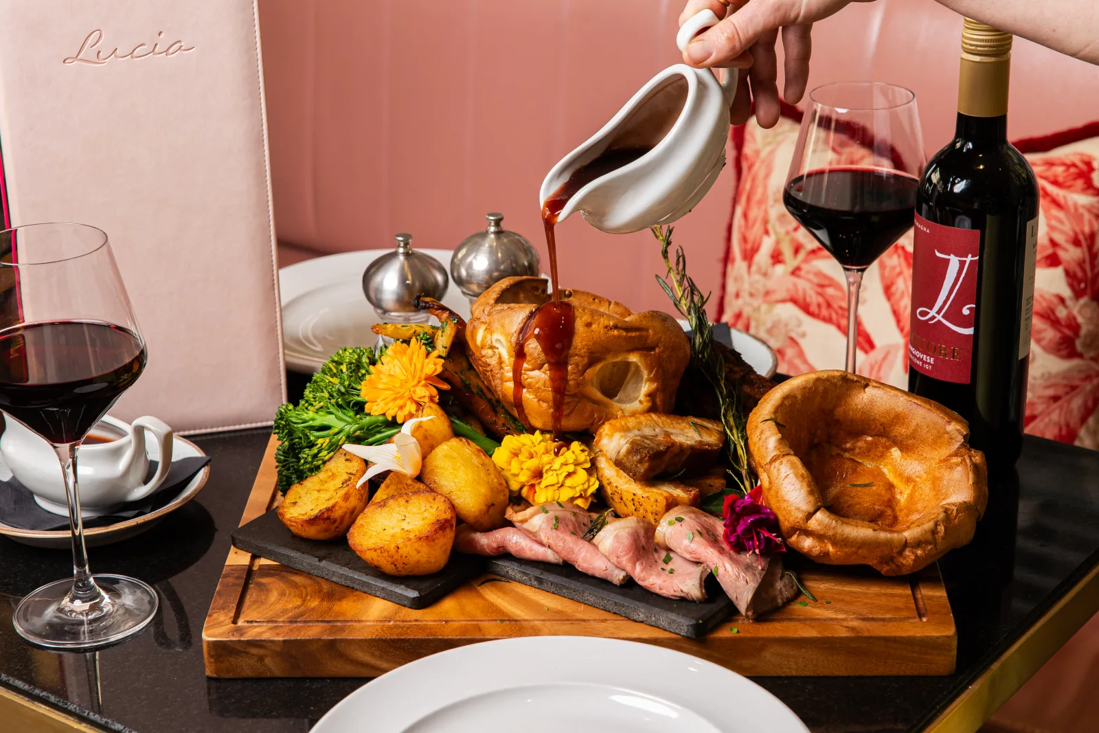 Elegant fine dining presentation at Lucia restaurant featuring roasted poultry and Yorkshire puddings on a wooden board, garnished with roasted potatoes, fresh herbs and edible flowers, with red wine being poured from a white gravy boat, accompanied by red wine in glasses and a bottle, set against a warm pink backdrop with table settings and menu visible.