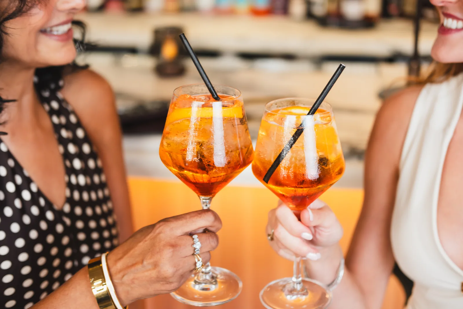 Two people holding colourful orange cocktails with ice and black straws at a beach venue, with blurred beach and other beachgoers visible in the background.