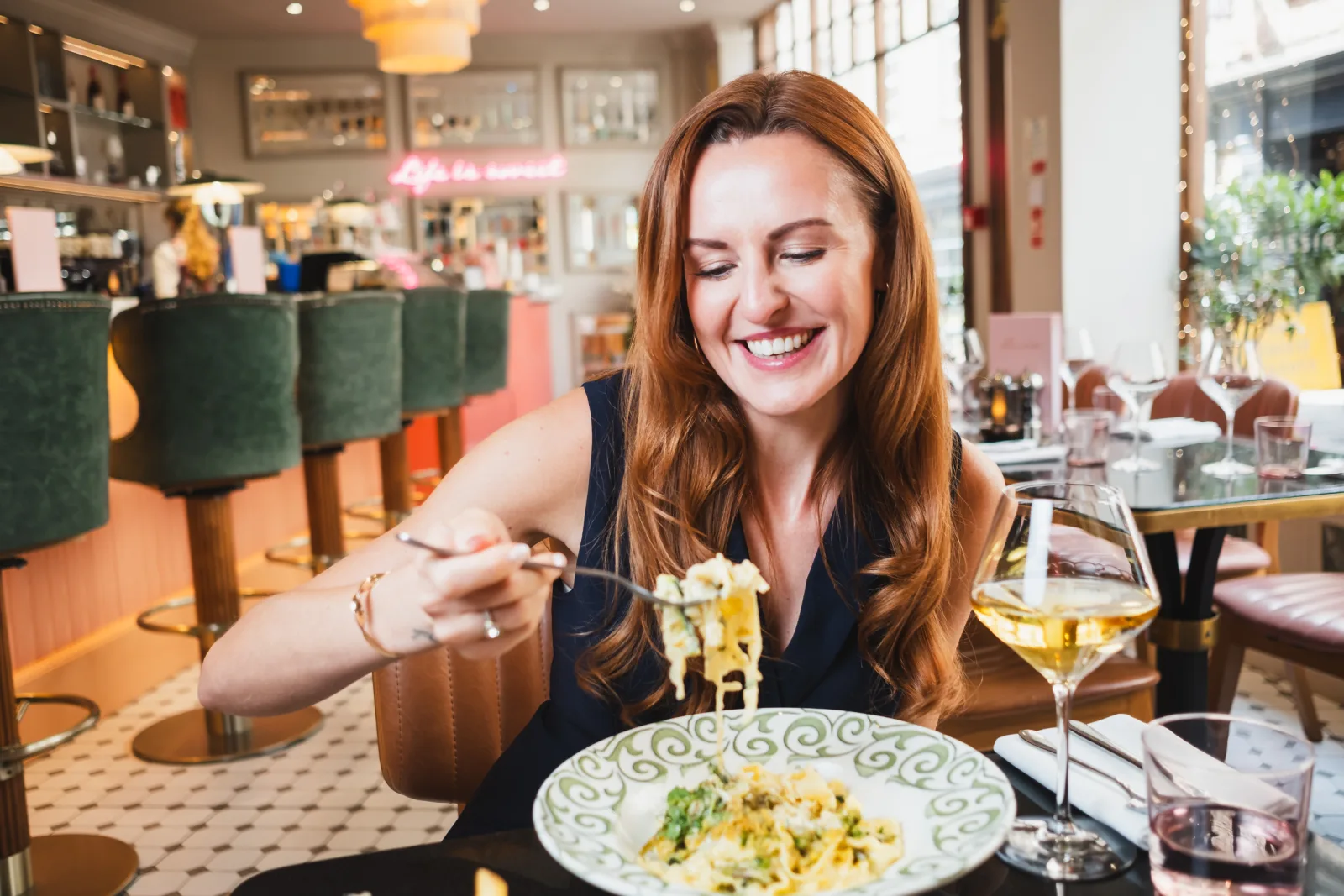 Person with reddish-brown hair dining at an upscale restaurant, smiling whilst eating pasta from a decorative plate, with a glass of white wine beside them and green bar stools and modern interior visible in the background.