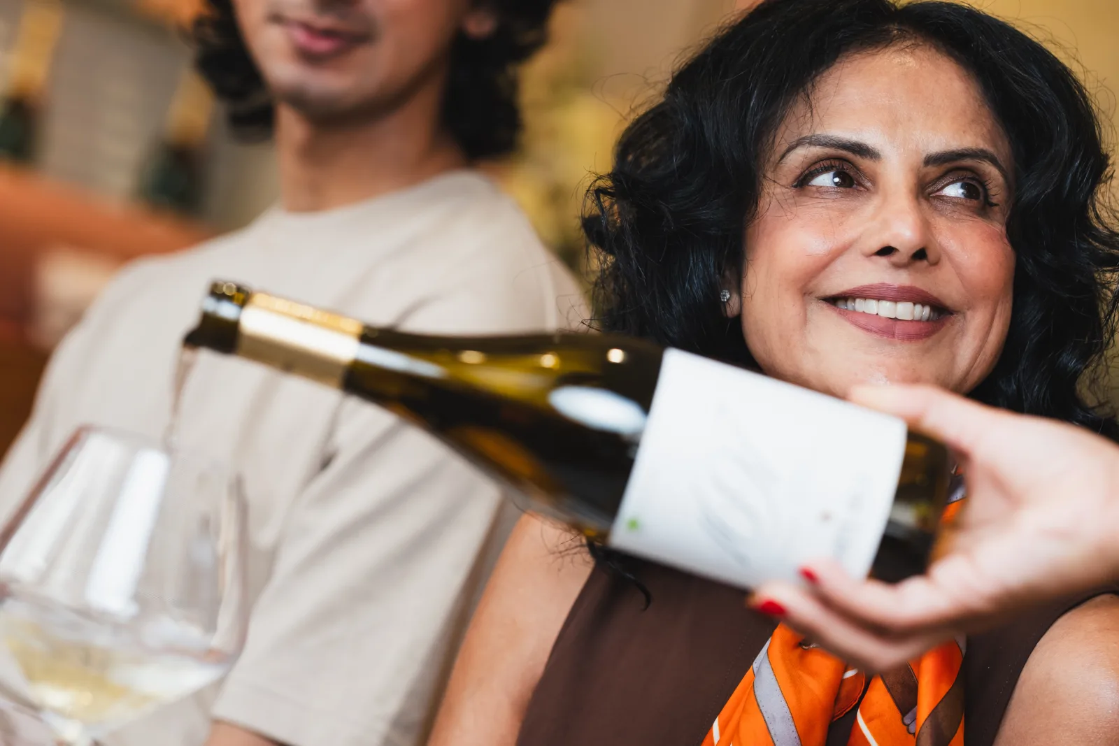 Person in orange top holding a wine bottle and white label, smiling at a social gathering with another person blurred in the background wearing cream-coloured clothing.