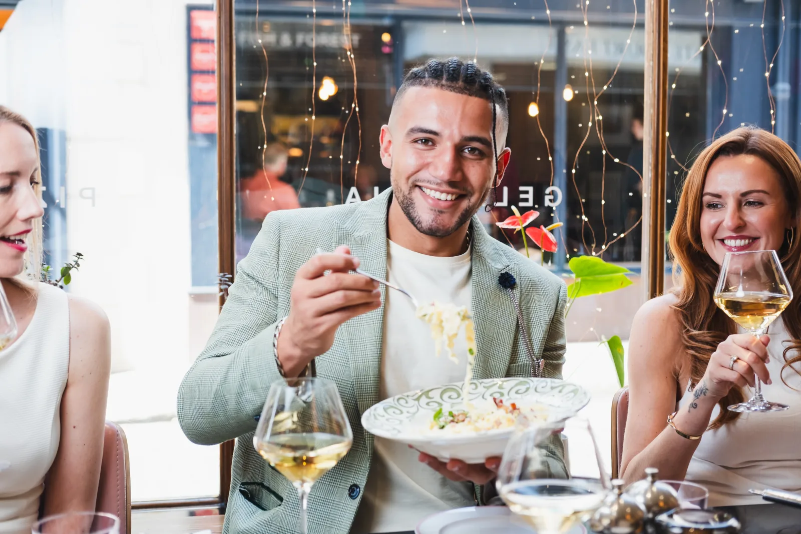 Three people dining at an upscale restaurant with festive string lights and decorations visible on the window behind them. The man in the centre, wearing a light grey blazer, is smiling whilst holding a plate of food. A woman on the left in a white top and a woman on the right holding a glass of white wine are also smiling. The table has wine glasses and plated dishes in front of them.