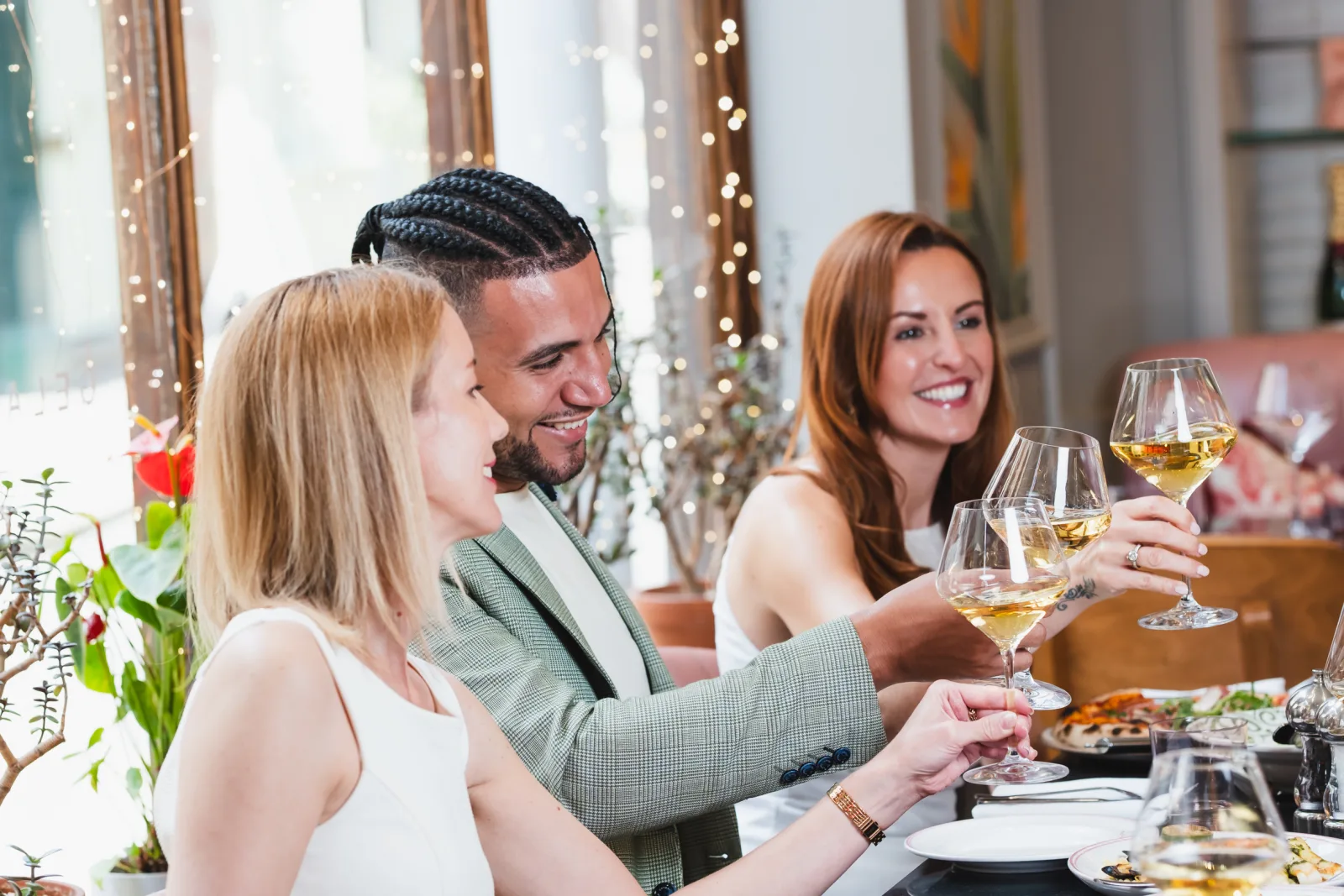 Three people enjoying drinks at an elegant dinner party with festive string lights, decorative curtains and floral arrangements in the background