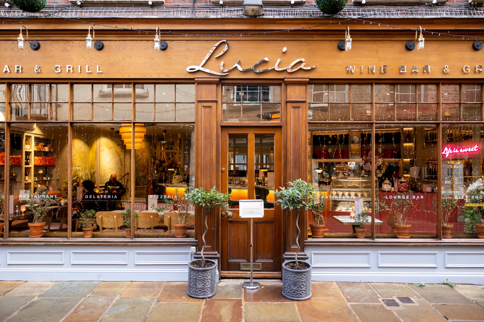 Lucia wine bar and grill storefront with wooden facade, large windows displaying restaurant and gelateria sections, potted plants flanking the entrance, and warm interior lighting