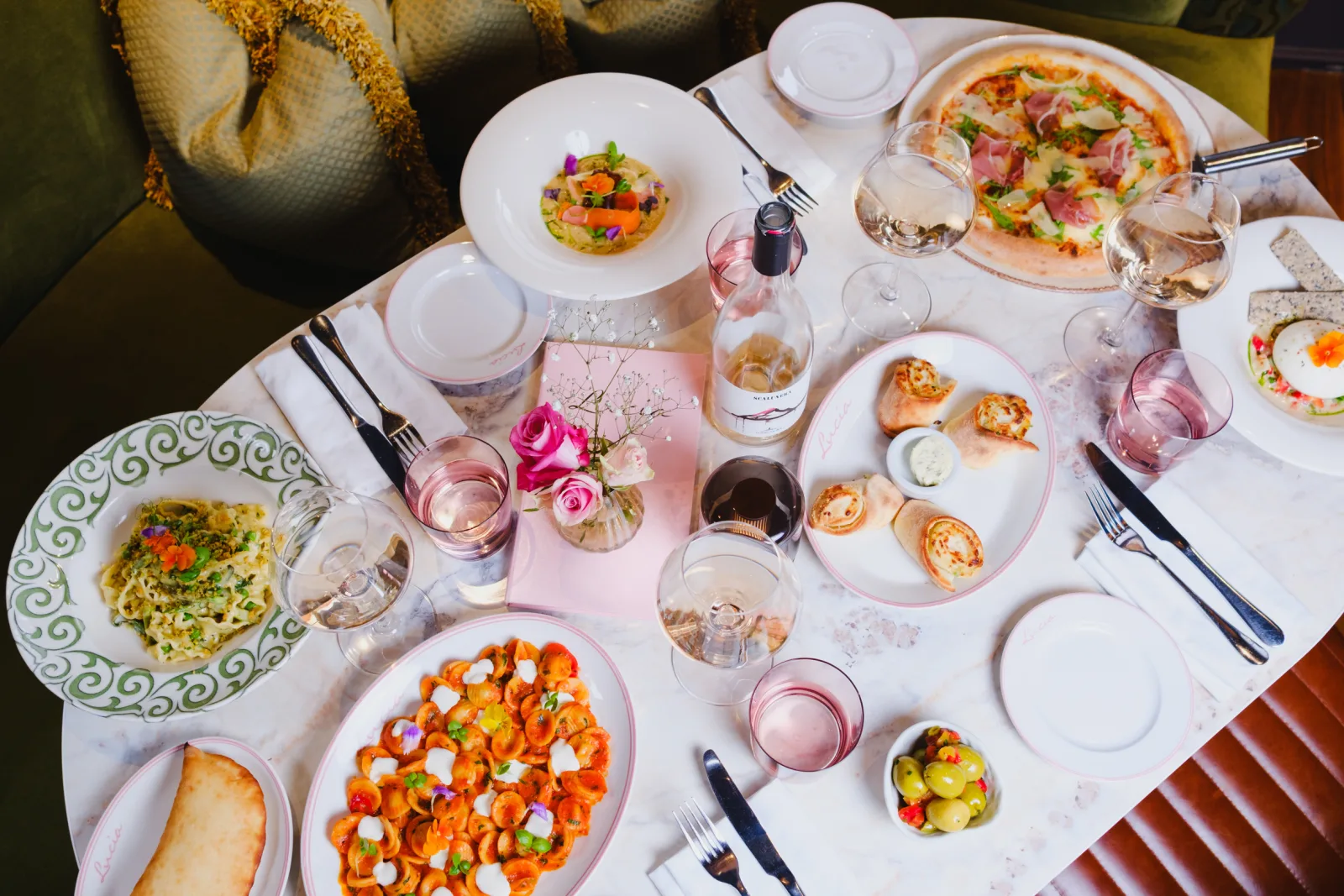 Overhead view of an elegantly set dining table with multiple colourful dishes including pizza, pasta, and appetisers, accompanied by glasses of rosé wine, fresh pink roses in a centrepiece, and place settings with cutlery and napkins.