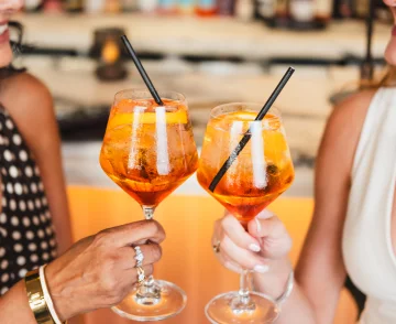 Two people holding colourful orange cocktails with ice and black straws at a beach venue, with blurred beach and other beachgoers visible in the background.