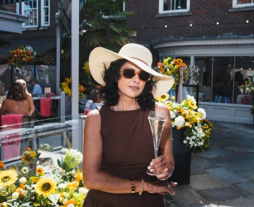 Person wearing a wide-brimmed straw hat and sunglasses holding a champagne flute at an outdoor garden party surrounded by yellow and white flowers, with brick buildings and other guests visible in the background.