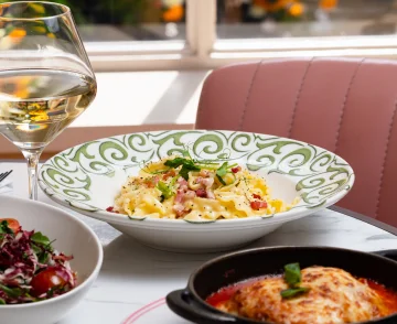 Fine dining table setting with a glass of white wine, a decorative plate of pasta carbonara with ham and green onions, a side salad with beetroot, and a black skillet dish, positioned near a pink quilted booth seat by a sunlit window with blurred garden background
