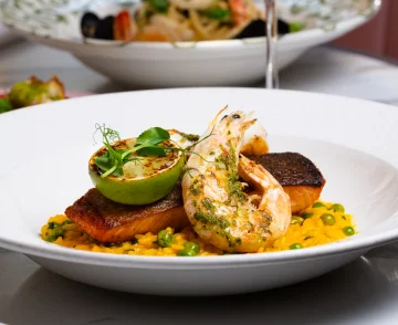 Fine dining plated dish featuring seared prawns and seared fish fillet with herb garnish, served on a bed of yellow corn puree with peas, topped with a halved grilled pear and microgreens, presented on a white plate with additional dishes visible in the blurred background