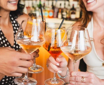 Two women smiling and toasting with glasses of amber-coloured cocktails with black straws at a bar, with shelves of bottles blurred in the background.