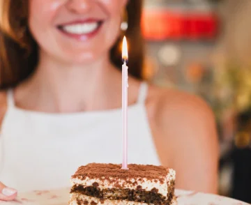 Smiling person with reddish-brown hair behind a layered chocolate and cream cake with a lit pink candle on top