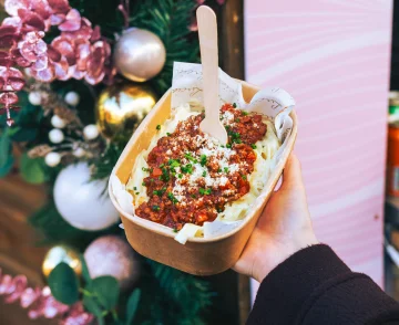 Hand holding a cardboard bowl of pasta topped with red sauce, grated cheese and green garnish with a wooden spoon, displayed in front of a festive Christmas decoration featuring pink flowers, baubles and evergreen foliage against a pink background.