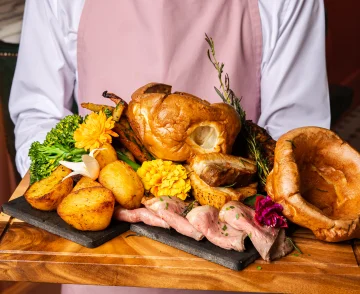 Chef in pink apron and white shirt holding a slate board displaying roasted duck with potatoes, corn, sliced ham, fresh herbs and edible flowers in a kitchen setting