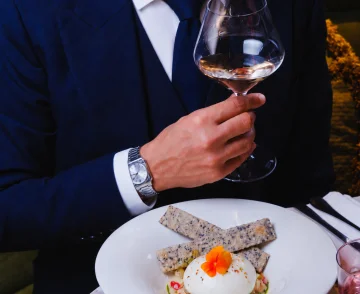 Man in navy suit and tie holding a glass of whiskey whilst seated at a table with a plated dish of poached egg topped with an orange flower and seeded crackers, wearing a watch and smiling.