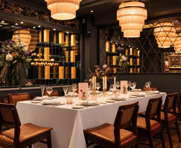 Elegant private dining room with a white tablecloth-covered table set for formal dinner, flanked by mid-century modern chairs with copper-coloured upholstery, ornate pendant lights overhead, decorative floral centrepieces, wine glasses and place settings, with dark walls featuring ornamental panels and illuminated shelving displaying bottles in the background.