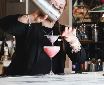 Bartender pouring a cocktail from a shaker into a pink drink at a marble-topped bar counter, wearing a black blazer and silver jewellery, with shelves of bottles and bar equipment visible in the background.