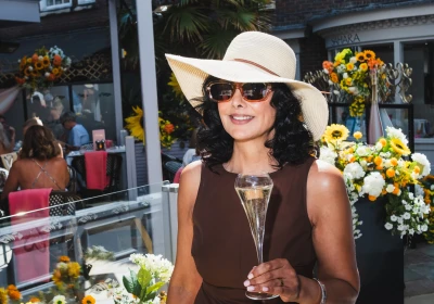 Person wearing a white wide-brimmed hat and sunglasses holds a champagne glass at an outdoor garden party surrounded by yellow and white flowers, with striped awning and brick buildings in the background.