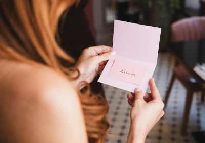 Person holding open a pale pink greeting card or invitation with "Lucia" written in cursive script inside, with blurred interior background visible behind them.
