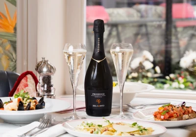 Elegant fine dining table setting with Bel Canto Prosecco bottle centred between two flute glasses filled with sparkling wine, surrounded by gourmet dishes including seafood and bruschetta, with white flowers and city views visible through large windows in the background.