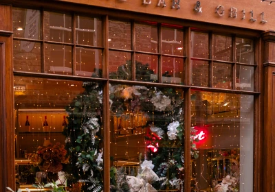 Wine bar and grill storefront with ornate wooden fascia, large multi-paned windows decorated with festive greenery and white flowers, string lights and decorative brass wall sconces above, red brick building visible at top, potted plants at street level.