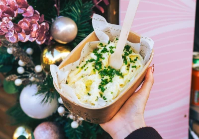 Person holding a cardboard takeaway container of creamy pasta garnished with fresh herbs and olive oil, positioned in front of festive Christmas decorations including pink baubles, evergreen branches and dried eucalyptus against a pink striped background.