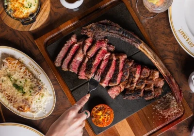 Overhead view of a wooden table set with sliced rare steak on a black cutting board, accompanied by various side dishes including salad, potatoes, and pickles, with a person's hand reaching toward the steak and glasses of wine and water visible around the spread.