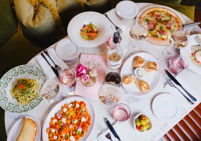 Overhead view of an elegantly set dining table with multiple colourful dishes including pizza, pasta, and appetisers, accompanied by glasses of rosé wine, fresh pink roses in a centrepiece, and place settings with cutlery and napkins.
