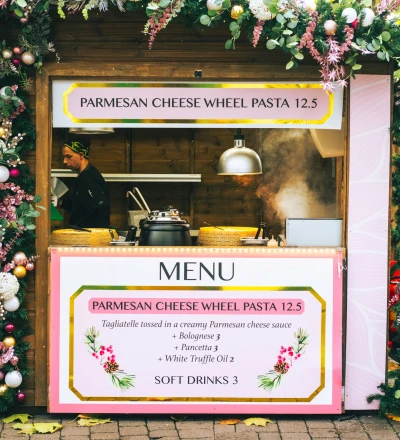 Decorated food stall selling Parmesan cheese wheel pasta, adorned with pink and white floral garlands, white baubles, and gold ornaments hanging from a wooden frame, with a menu displayed below showing pasta and soft drinks options.