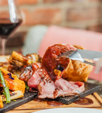 Plated gourmet meat dish with a knife cutting into a glazed red meat portion, accompanied by roasted vegetables and edible flowers on a slate board, with a wine glass and brick background