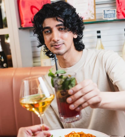 Person with black wavy hair wearing a beige t-shirt holding a dark cocktail with mint garnish at a dining table, with another person holding a glass of white wine beside a plate of food with orange-coloured fish and lemon slices, shelving with red heart decorations visible in the background.