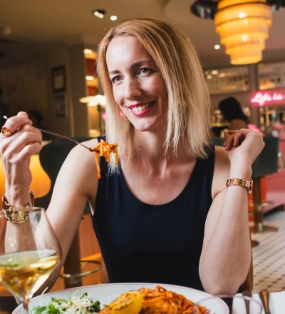 Person with reddish-blonde hair wearing a black sleeveless top and gold bracelets, smiling whilst eating pasta with a fork at a restaurant table with a wine glass and plated food, with warm lighting and pink neon signage visible in the background.