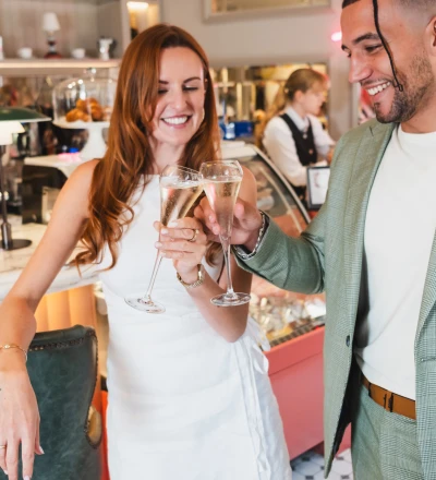 Two people toasting with champagne glasses at an upscale bar or restaurant, both smiling and wearing formal attire, with bartenders and interior décor visible in the background.