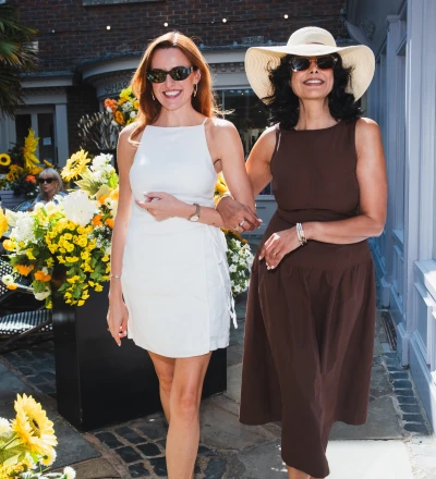 Two people walking through a sunny courtyard wearing sunglasses, surrounded by yellow and white flowers, with a brick building and white-framed windows in the background.