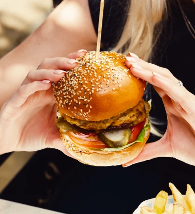 Person holding a sesame seed burger with a fried patty, pickles, tomato and lettuce, with a wooden pick through the top, wearing a black shirt and red lipstick, with French fries visible in the foreground