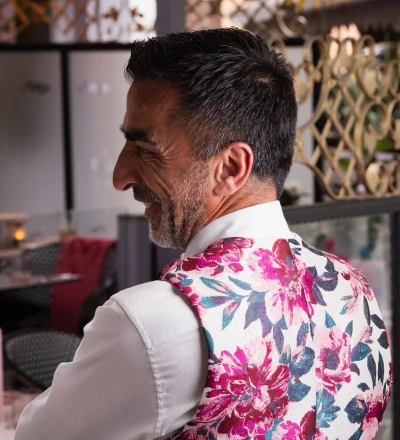 Person with dark hair and beard wearing a white shirt and vibrant pink and blue floral patterned waistcoat, shown in profile view under a blue and white striped awning in what appears to be a Mediterranean-style interior space.