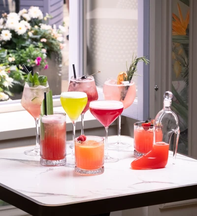 Colourful cocktails and mocktails arranged on a white marble table by a window, featuring pink, red, yellow and orange drinks garnished with fresh herbs and citrus, with potted flowers visible outside