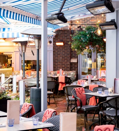 Outdoor restaurant patio with blue and white striped retractable awning, black and white patterned chairs, coral napkins and table settings, infrared heaters mounted overhead, brick building backdrop with climbing ivy, and warm evening lighting.