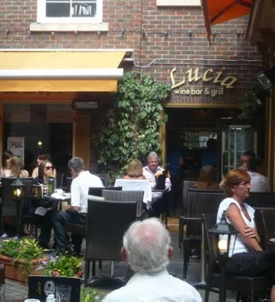 Outdoor restaurant terrace with diners seated at black tables under a yellow awning, ivy-covered storefront, brick building facade with Lucia wine bar and grill signage visible.
