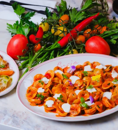 Fine dining table setting with two white plates with pink rims containing plated dishes, featuring roasted orange vegetables topped with white cream and edible flowers, alongside fresh tomatoes, red chillies, yellow peppers and parsley garnish in the centre, with cutlery and a glass of dark liquid visible.