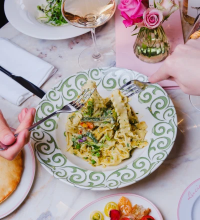 Elegant dining table with person's hands holding fork over decorative green and white plate of pasta dish, wine glass with white wine, pink roses and baby's breath in glass vase, and colourful appetiser plate with edible flowers below.