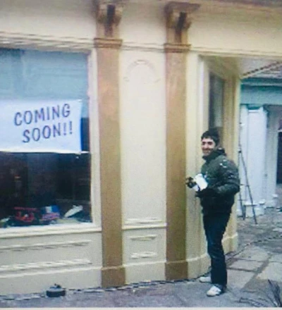 Person standing in front of a storefront with "cia COMING SOON!!" written on the window, featuring cream-coloured facade with brown wooden pillars and residential buildings visible in the background.