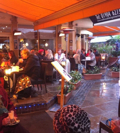Bustling covered outdoor café area at dusk with numerous patrons seated at tables under orange canopy, warm lighting, potted plants, and a café sign visible in the background.