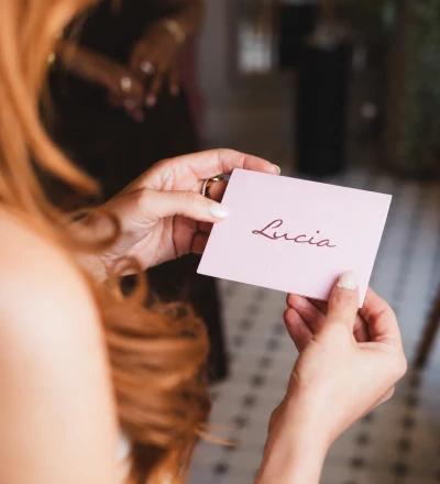 Person holding a pale pink business card with the name "Lucia" written in burgundy script handwriting, wearing a mustard-coloured top with rings on their fingers
