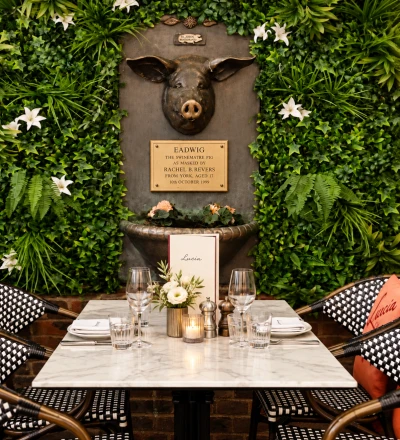 Dining table set for two with marble top and black and white patterned chairs, positioned in front of a lush green living wall featuring white flowers. A bronze pig head sculpture with plaque reading "Eadwig the Swinematre Pig" is mounted on a slate background, with decorative bronze ornaments and a flower-filled urn below it.