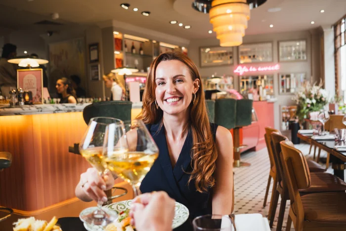 Person with reddish-brown hair wearing a dark blue sleeveless top, holding a glass of white wine whilst smiling at a restaurant bar with warm lighting, comfortable seating, and shelves of bottles in the background.